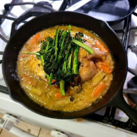 Hearty braised chicken and carrots topped with vibrant broccolini in a cast-iron skillet on a gas stovetop.
