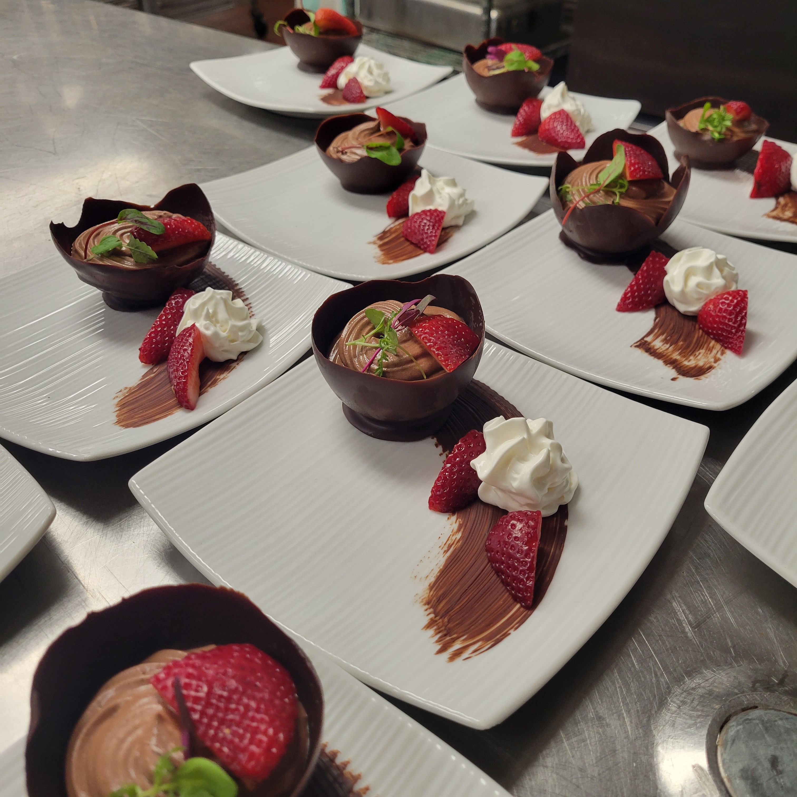 Plated gourmet desserts: chocolate mousse served in chocolate cups with fresh strawberry slices, whipped cream and chocolate smear on white square plates in a professional kitchen