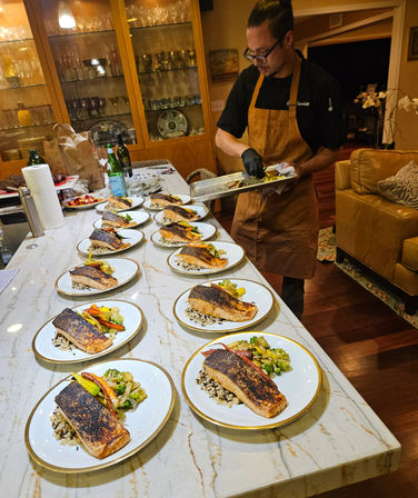 Chef in a home kitchen plating rows of seared salmon fillets on wild rice with sautéed vegetables on white gold-rim plates along a marble island for a private dinner service.