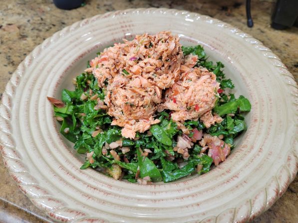 Three scoops of shredded salmon atop chopped kale and whole-grain salad on a decorative ceramic plate on a kitchen countertop.