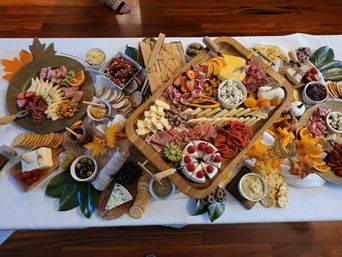 Overhead view of an autumn-themed charcuterie grazing board on a white tablecloth — wooden trays piled with sliced cheeses, cured meats, crackers, olives, nuts, fruit (strawberries, kiwi, orange), dips and decorative fall leaves.