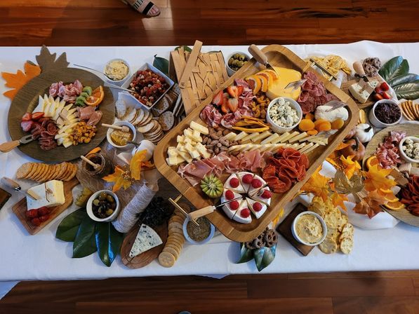 Overhead view of an autumn-themed charcuterie grazing board on a white tablecloth — wooden trays piled with sliced cheeses, cured meats, crackers, olives, nuts, fruit (strawberries, kiwi, orange), dips and decorative fall leaves.