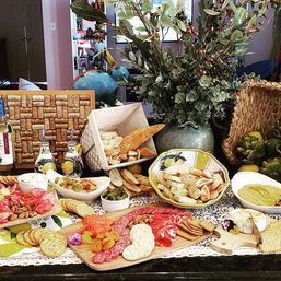 Inviting home charcuterie spread on a kitchen counter with sliced baguette and bread basket, assorted crackers, cured meats and cheeses, nuts, olives, pickles, dried fruit and dips, styled with a large vase of greenery and woven baskets