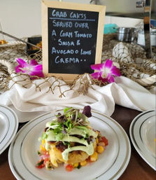 Plated crab cake appetizer topped with microgreens and avocado-lime crema, served on corn and tomato salsa with a chalkboard sign and purple orchid garnishes in the background.