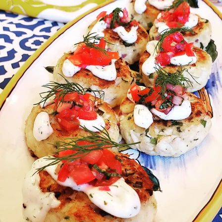 Close-up of golden pan-seared seafood cakes on an oval platter topped with creamy herb sauce, bright tomato salsa and fresh dill sprigs — a colorful seafood appetizer perfect for parties.