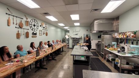 Chef demonstrating in a bright culinary studio with students seated along a long communal table set for a hands-on cooking class.
