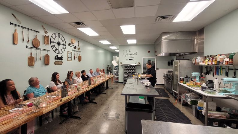 Chef demonstrating in a bright culinary studio with students seated along a long communal table set for a hands-on cooking class.