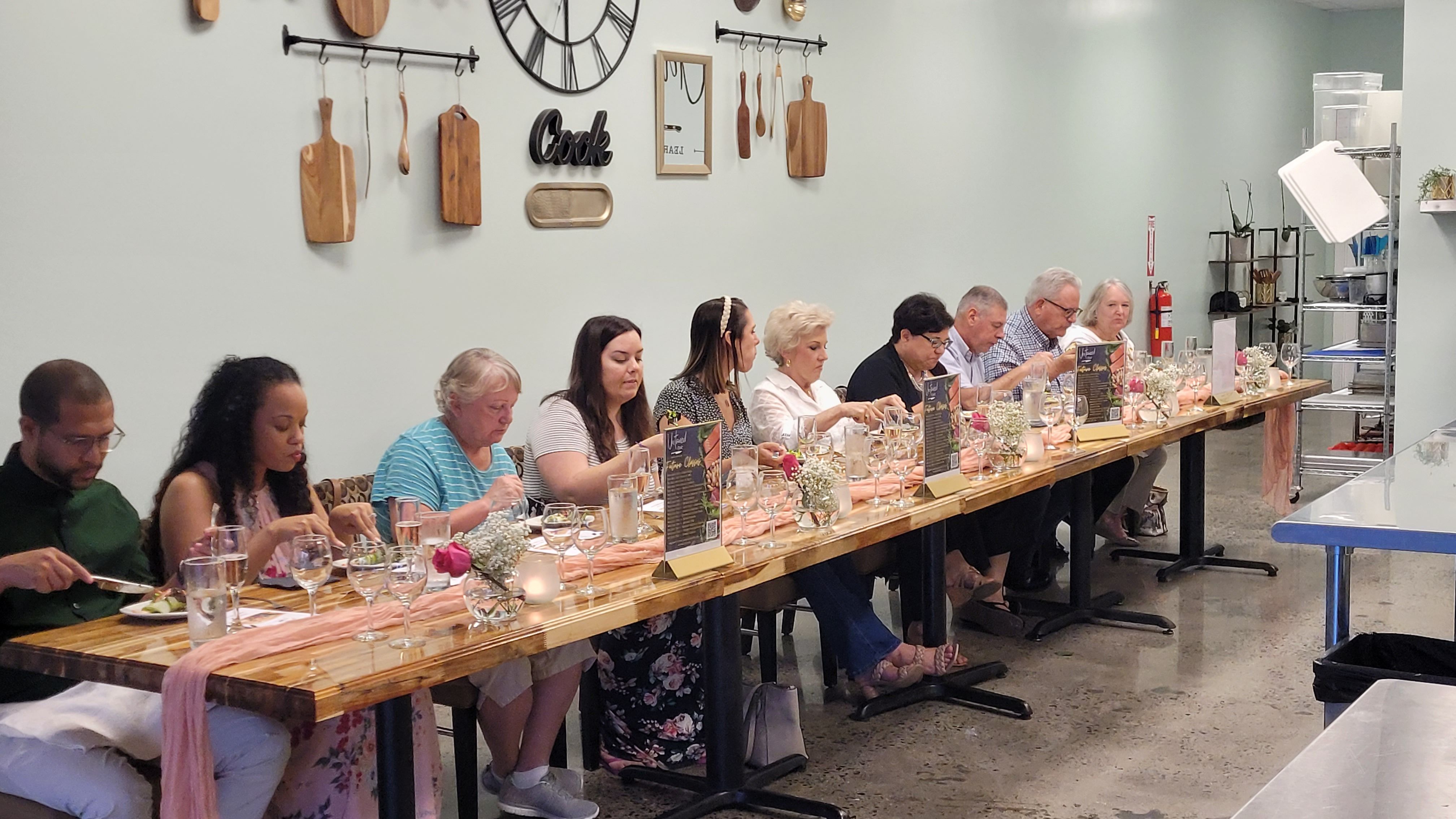 Communal tasting dinner in a bright cooking studio: guests seated at a long wooden table with wine glasses, menus, floral centerpieces and wall-mounted cutting boards and decorative clock.