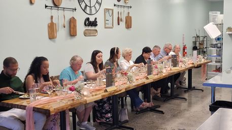 Communal tasting dinner in a bright cooking studio: guests seated at a long wooden table with wine glasses, menus, floral centerpieces and wall-mounted cutting boards and decorative clock.