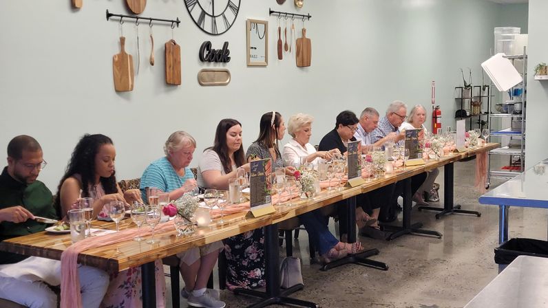 Communal tasting dinner in a bright cooking studio: guests seated at a long wooden table with wine glasses, menus, floral centerpieces and wall-mounted cutting boards and decorative clock.