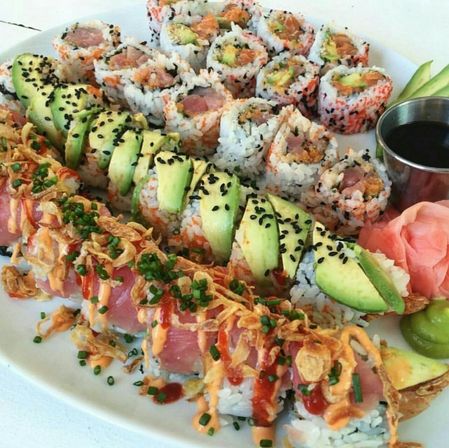 Colorful sushi platter on a white plate — avocado-topped rolls and tuna rolls with black sesame, crispy fried onions, spicy mayo drizzle, soy sauce, pickled ginger and wasabi