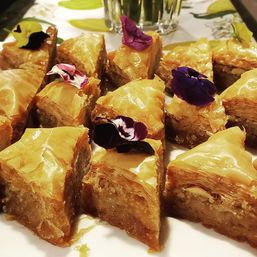 Close-up of golden triangular baklava slices with flaky phyllo, syrupy nut filling and decorative edible pansy flowers — Mediterranean honey pastry dessert on a plate.