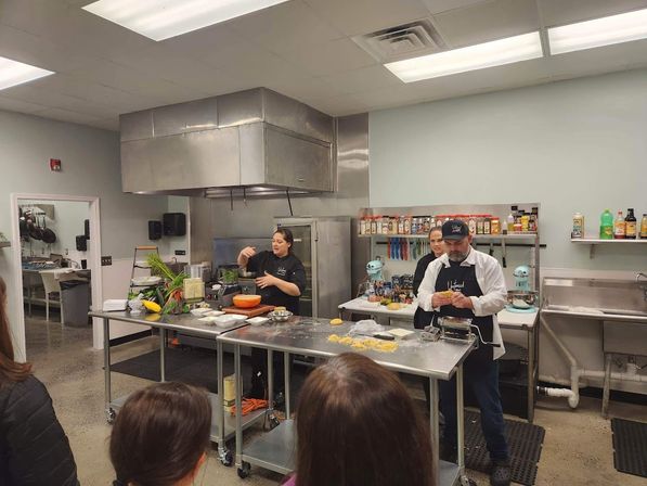 Hands-on cooking class in a commercial kitchen with instructors demonstrating fresh pasta making at stainless-steel prep tables, pasta roller, stand mixers and shelves of spices while a small audience watches.