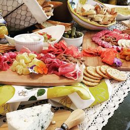 Party-ready gourmet charcuterie spread on wooden boards — blue cheese wedge, marinated mozzarella, assorted cured meats, crackers, pickles and sliced bread on a decorative tablecloth