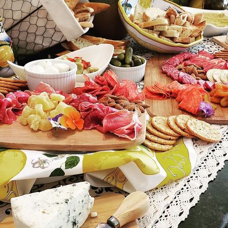 Party-ready gourmet charcuterie spread on wooden boards — blue cheese wedge, marinated mozzarella, assorted cured meats, crackers, pickles and sliced bread on a decorative tablecloth