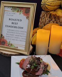 Plated roasted beef tenderloin with demi-glace, microgreens, whipped potatoes and roasted cherry tomato on a white plate next to a gold-framed "Roasted Beef Tenderloin" menu sign, glowing candles and autumn gourds