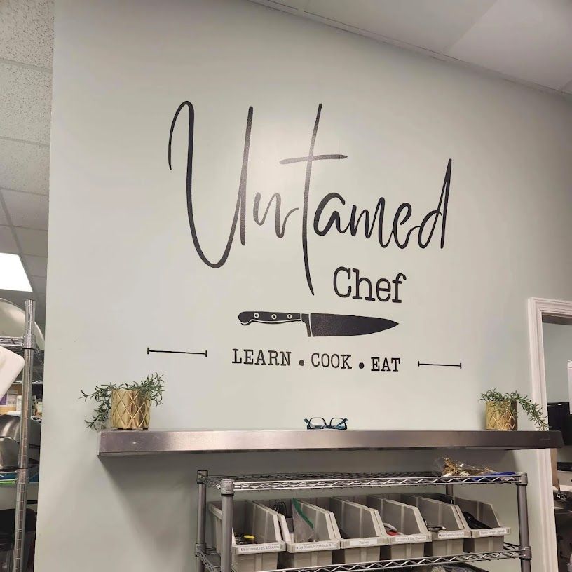 Decorative wall decal in a commercial kitchen: large script lettering with a chef’s knife graphic and the slogan LEARN • COOK • EAT above a stainless shelf with labeled bins and small potted plants.