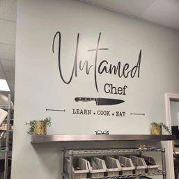 Decorative wall decal in a commercial kitchen: large script lettering with a chef’s knife graphic and the slogan LEARN • COOK • EAT above a stainless shelf with labeled bins and small potted plants.