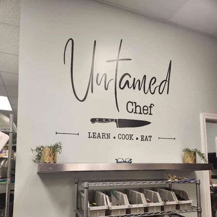 Decorative wall decal in a commercial kitchen: large script lettering with a chef’s knife graphic and the slogan LEARN • COOK • EAT above a stainless shelf with labeled bins and small potted plants.