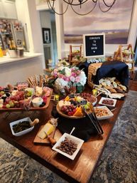 Festive bridal shower charcuterie spread on a wooden dining table in a home, featuring assorted cheeses, crackers, cured meats, grapes, nuts, dips, floral centerpiece and a letterboard that reads 'Congrats to the bride'.