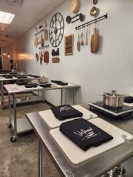 Interior of a modern cooking studio with rows of stainless-steel prep tables, white cutting boards topped with black aprons, induction burners and pots; wall decorated with wooden utensils, cutting boards and a large metal clock.
