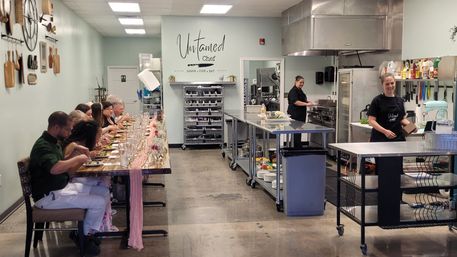 Guests seated at a long communal table enjoying plated dishes in a bright culinary studio with stainless-steel prep islands and chefs cooking