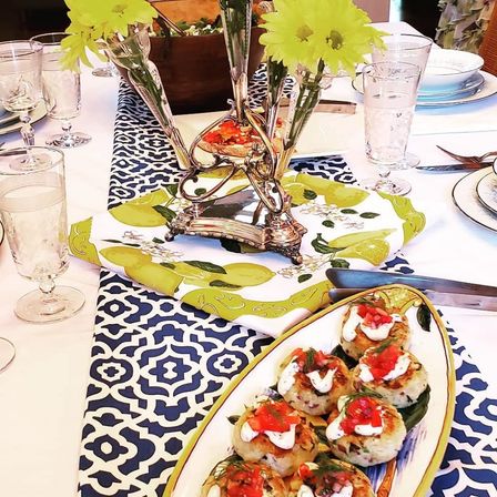 Sunny spring tablescape with blue geometric runner, lemon-print napkin and silver vase of yellow flowers; oval platter of bite-sized savory appetizers topped with cream and diced tomato, surrounded by glassware and white plates.