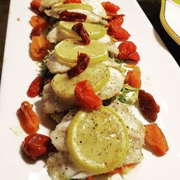 Zesty lemon-topped baked white fish fillets on a long white platter, drizzled with olive oil and herbs and arranged with roasted cherry tomatoes and shredded greens