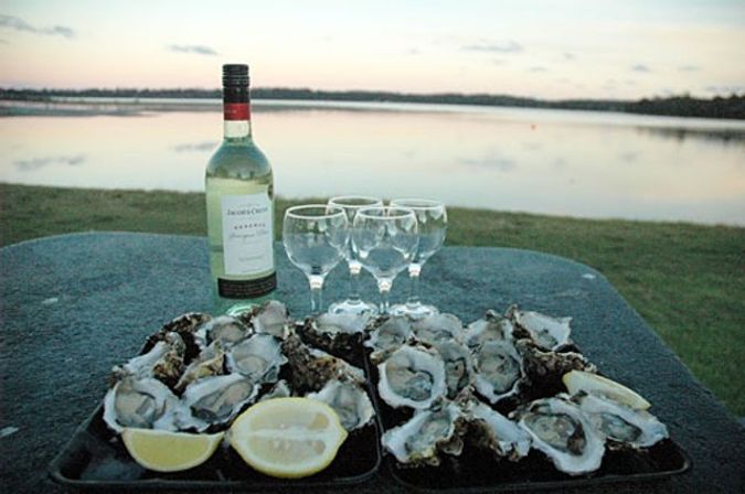 Lakeside sunset spread: trays of freshly shucked oysters with lemon wedges, a bottle of white wine and four wine glasses on a picnic table by calm water.