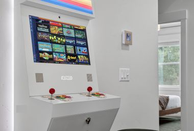 White retro arcade cabinet with red joysticks and colorful classic game-selection screen in a bright modern home hallway beside a bedroom window