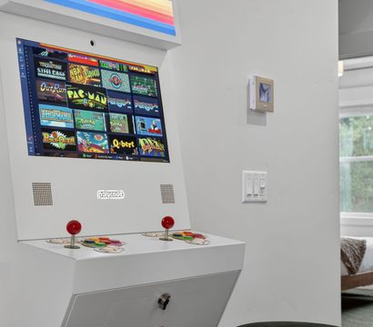 White retro arcade cabinet with red joysticks and colorful classic game-selection screen in a bright modern home hallway beside a bedroom window