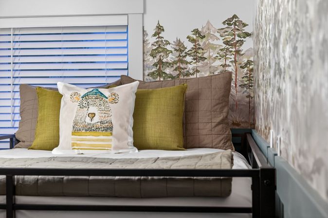 Cozy nature-themed bed with a playful bear throw pillow, olive and taupe cushions, quilted bedding, pine forest wall mural and white window blinds.