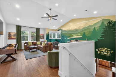 Bright loft living room with brown leather sectional, green throw pillows, shuffleboard table and hardwood floors, vaulted ceiling with fan, corner dining nook and large stylized mountain mural — vacation rental vibe in Sunriver, Oregon.