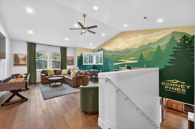 Bright loft living room with brown leather sectional, green throw pillows, shuffleboard table and hardwood floors, vaulted ceiling with fan, corner dining nook and large stylized mountain mural — vacation rental vibe in Sunriver, Oregon.