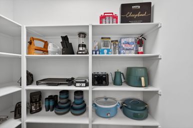 Organized walk-in pantry with white shelves displaying small kitchen appliances and cookware, including a teal air fryer, toaster, kettle, blender, coffee maker, electric griddle, stacked blue-gray bowls and a slow cooker.