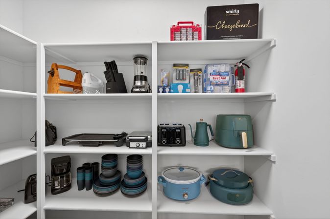 Organized walk-in pantry with white shelves displaying small kitchen appliances and cookware, including a teal air fryer, toaster, kettle, blender, coffee maker, electric griddle, stacked blue-gray bowls and a slow cooker.