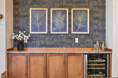 Cozy home bar interior with granite countertop and wooden cabinets, built-in wine fridge, dark textured wallpaper, three framed botanical pine prints, black vase of white blooms and a small cocktail set.