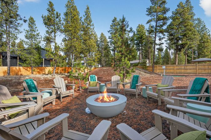 Round outdoor fire pit with dancing flames surrounded by gray Adirondack chairs draped with teal blankets in a pine‑wooded backyard seating area