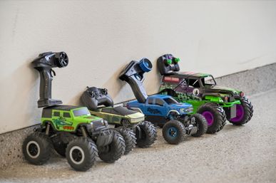 Row of colorful remote-controlled (RC) monster trucks with handheld controllers lined up against a white wall on a concrete garage floor.
