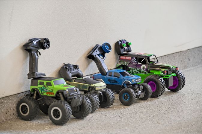 Row of colorful remote-controlled (RC) monster trucks with handheld controllers lined up against a white wall on a concrete garage floor.