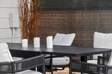 Cozy modern backyard patio dining setup: black metal table with three white pillar candles and gray cushioned chairs by a textured privacy screen.