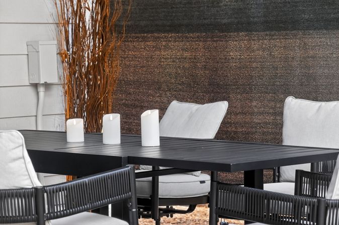 Cozy modern backyard patio dining setup: black metal table with three white pillar candles and gray cushioned chairs by a textured privacy screen.