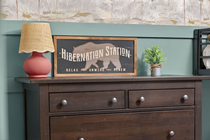 Cozy farmhouse vignette of a dark wood dresser topped with a pink ceramic lamp, Hibernation Station bear sign, and small potted plant against a teal shiplap wall.