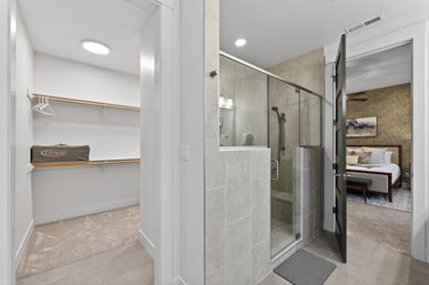 Bright modern en-suite bathroom in a contemporary home with a glass-enclosed gray-tiled shower, an adjacent carpeted walk-in closet with wooden hanging rods, and a doorway revealing a styled bedroom with a patterned accent wall and bed.