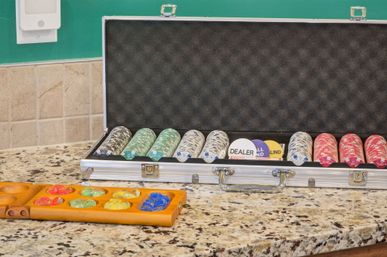 Open aluminum poker chip case with stacked red, white and green chips and dealer/blind buttons beside a wooden mancala board with colorful glass stones on a speckled granite countertop and tiled backsplash, ready for game night.