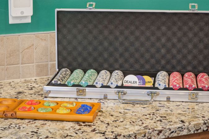 Open aluminum poker chip case with stacked red, white and green chips and dealer/blind buttons beside a wooden mancala board with colorful glass stones on a speckled granite countertop and tiled backsplash, ready for game night.