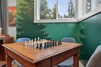 Cozy game nook with a wooden table featuring an inlaid chessboard and black and white pieces, gray chairs, green forest mural walls and a window overlooking pine trees