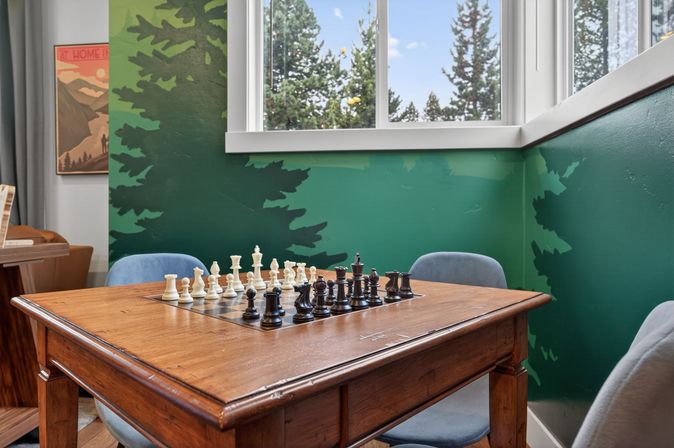 Cozy game nook with a wooden table featuring an inlaid chessboard and black and white pieces, gray chairs, green forest mural walls and a window overlooking pine trees