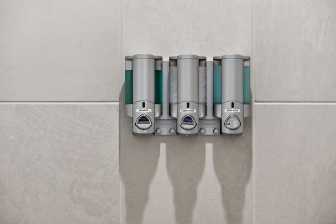 Triple wall-mounted dispensers labeled shampoo, conditioner and body wash on a light gray tiled shower wall — refillable bathroom shower station