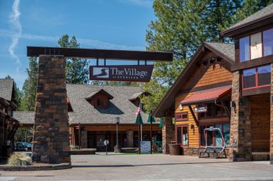 Rustic mountain village entrance with stone pillar arch, wooden lodges and shops, visitor information sign, repurposed chairlift bench, pine trees and blue sky